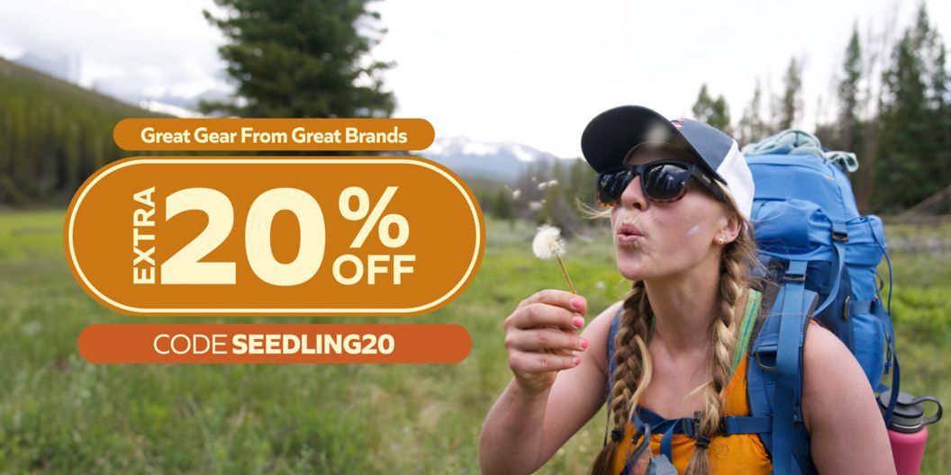 A woman backpacking in an alpine area blows on a dandelion. Text overlay reads Great Gear From Great Brands, Extra 20% off, Code SEEDLING20.