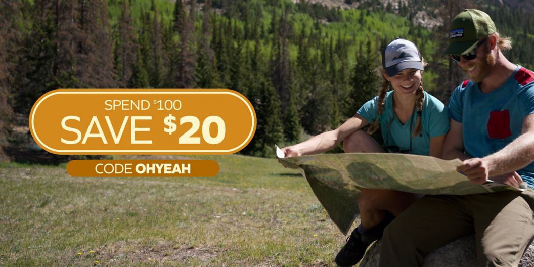 Two hikers in a mountain meadow look at a map together. Overlaid text reads, “Spend $100, save $20. Code OHYEAH” 