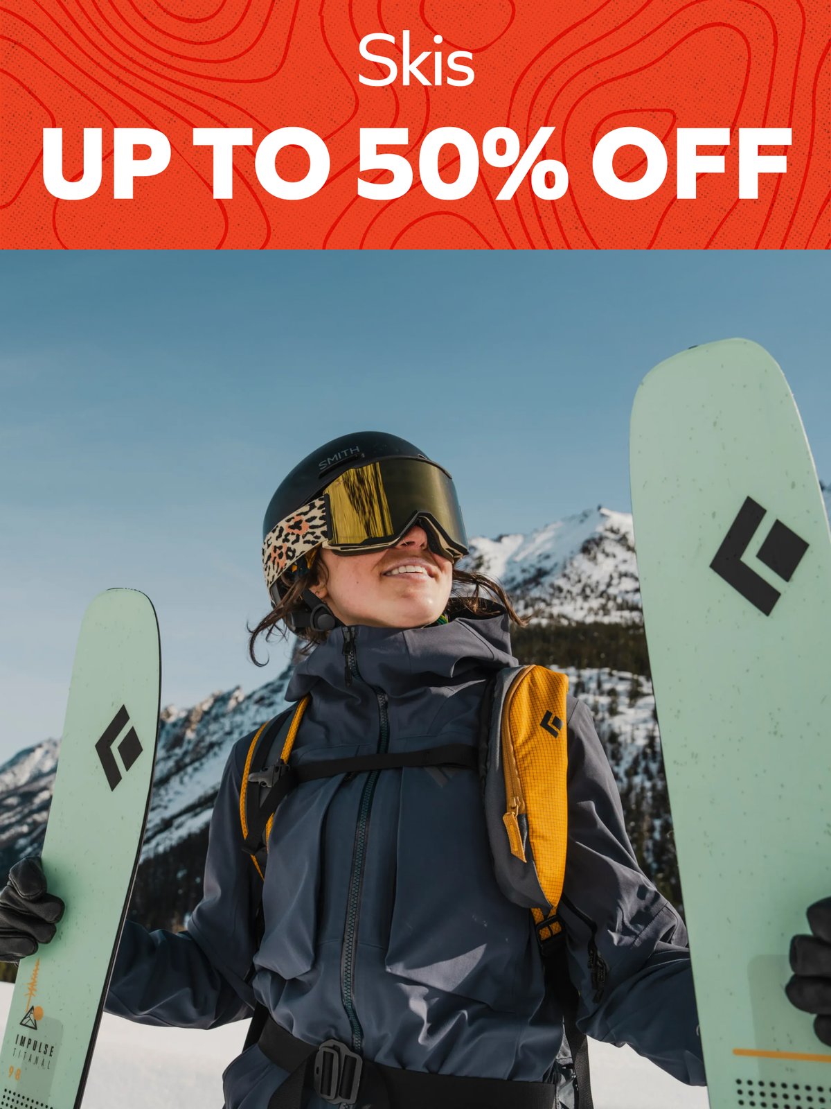 Skis up to 50% off — smiling skier in helmet and goggles holding a pair of skis in the snowy mountains.